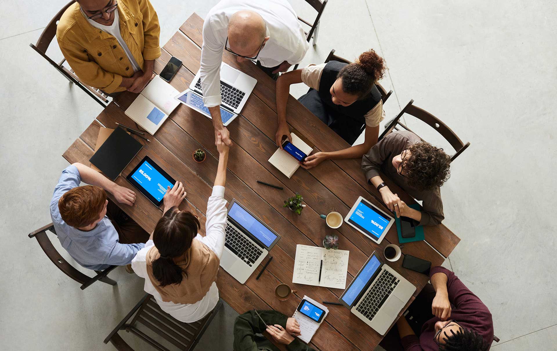 group of people sitting around a table, all have laptops. a man reaches across the table to shake a woman's hand.